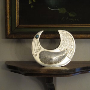 May include: A silver-colored, bird-shaped decorative object with an open crescent design. The bird has detailed wing and tail patterns, with a small blue gemstone accent. It sits on a wooden shelf, with a framed painting in the background.
