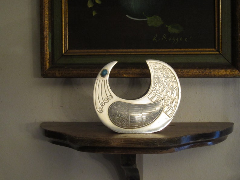 May include: A silver-colored, bird-shaped decorative object with an open crescent design. The bird has detailed wing and tail patterns, with a small blue gemstone accent. It sits on a wooden shelf, with a framed painting in the background.