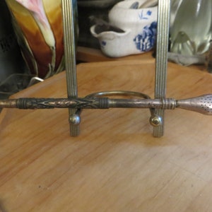 May include: A silver-toned metal pipe with a decorative design and a perforated mouthpiece. The pipe is displayed on a wooden stand.