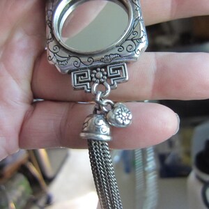 Pu&ograve; includere: Un specchietto compatto argentato e ornato con una cornice rettangolare e un piccolo specchio rotondo. Il compatto presenta motivi floreali e geometrici decorativi, un nappa e due piccoli ciondoli. Il compatto &egrave; tenuto in una mano.