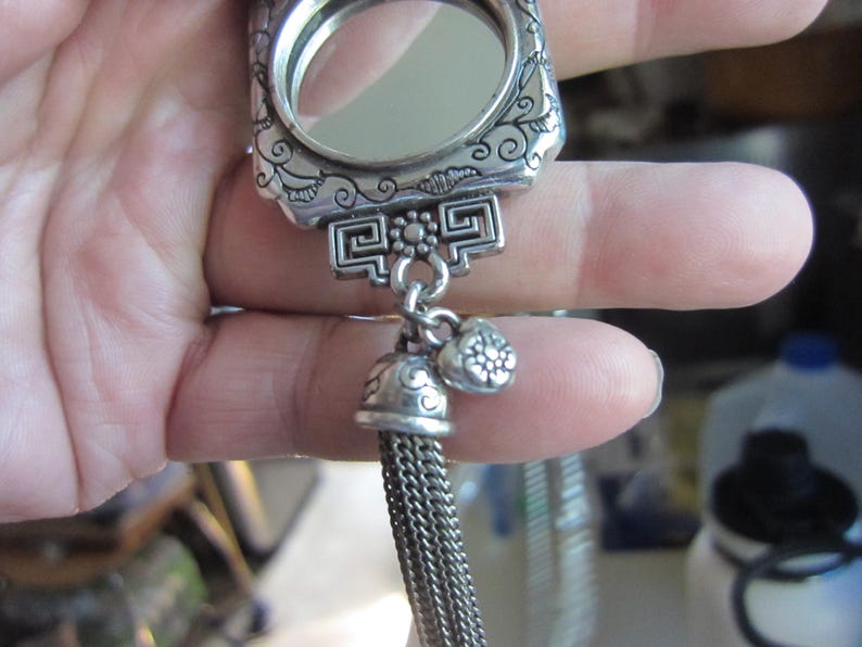 Pu&ograve; includere: Un specchietto compatto argentato e ornato con una cornice rettangolare e un piccolo specchio rotondo. Il compatto presenta motivi floreali e geometrici decorativi, un nappa e due piccoli ciondoli. Il compatto &egrave; tenuto in una mano.