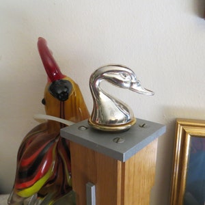 May include: A silver duck head bottle stopper with a gold base, mounted on a wooden post with a gray square base.