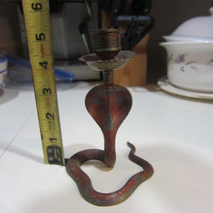 May include: A vintage cobra-shaped candle holder, primarily red with brass accents. The candle holder is approximately 5 inches tall. The cobra's head forms the candle cup, and the body coils along the base. The candle holder has a distressed finish.
