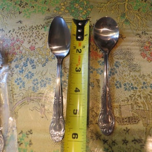 May include: Two silver spoons with ornate handles, measured against a yellow tape measure. The spoons are approximately 5 inches long.