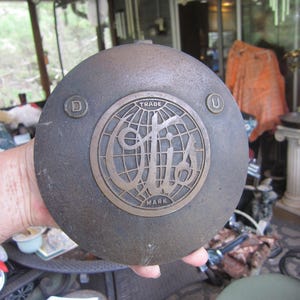 May include: A round, dark metal object with a bronze-colored emblem featuring a globe design and the text "TRADE MARK". The object has the letters "D" and "U" on either side of the emblem.