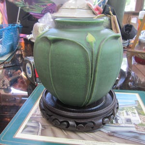 May include: A green ceramic vase with a floral design, resting on a dark wooden stand. The vase has a bulbous shape with a textured surface and a single yellow bud detail. The stand is round with carved detailing.