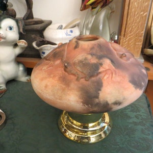 May include: A ceramic vase with a wide, rounded top and a gold-colored base. The vase has a reddish-brown color with darker brown accents and a textured surface. A ceramic dog figurine is in the background.