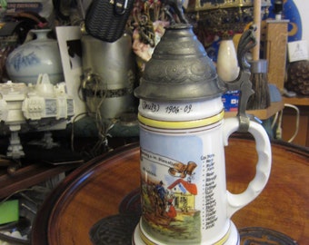 May include: A white ceramic beer stein with a pewter lid and a knight on horseback figurine. The stein has a colorful scene of people in a village and a list of German words. The stein is on a wooden tray.
