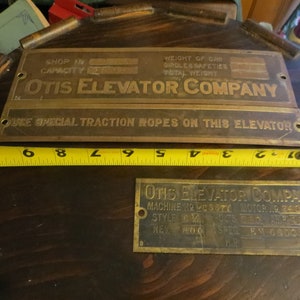 May include: Close-up of three brass plates with black lettering. The top plate reads "OTIS ELEVATOR COMPANY" and has the text "SHOP NO. CAPACITY" and "WEIGHT OF CAR, GIRGLES SAFETIES, TOTAL WEIGHT". The middle plate reads "USE SPECIAL TRACTION ROPES ON THIS ELEVATOR". The bottom plate reads "OTIS ELEVATOR COMPANY" and has the text "MACHINE NO. 12 MC 5771, MOTOR NO. 24 1724, STYLE GM, SPECS, REV. 1100,  R.M. 66003".
