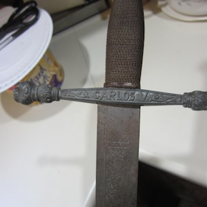 May include: Close-up of a sword with a textured brown handle and a silver crossguard. The crossguard has the inscription "CARLOS V" and decorative details. The blade shows etched patterns. A plate and scissors are in the background.