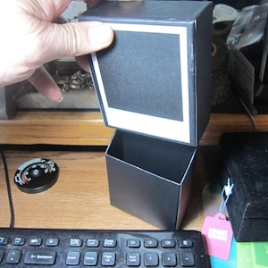 May include: A black box with a white frame around a black square. The box is open and the top part is sitting on top of the bottom part. The box is on a wooden surface.