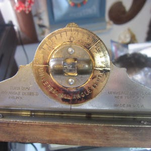 May include: A vintage Universal All Angle Level tool. The level has a brass dial and a silver-coloured metal base. The text "UNIVERSAL MFG. CO. NEW YORK, N.Y. MADE IN U.S.A." is visible. The tool is mounted on a wooden base.