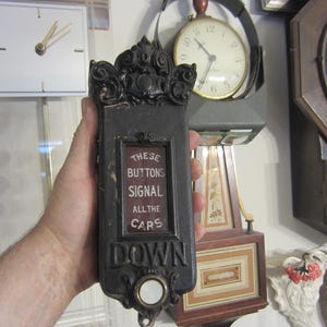 May include: An antique black elevator call button with ornate detailing. The rectangular button has a framed window with the text "THESE BUTTONS SIGNAL ALL THE CARS DOWN". A white button is at the bottom.
