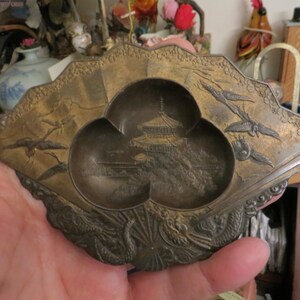 May include: A gold-coloured metal trinket dish with three compartments. The dish is decorated with a raised design of a pagoda and stylised birds. The dish is in the shape of a fan.