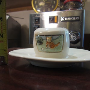 May include: White ceramic cup and saucer with a cat and a bird design. The cat is orange and white and the bird is green. The cup and saucer are decorated with gold trim.
