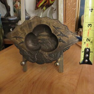 May include: A decorative brass tray with a fan-shaped design. The tray features a raised, three-dimensional image of a pagoda and a landscape. The tray is on a small, gold-coloured stand.
