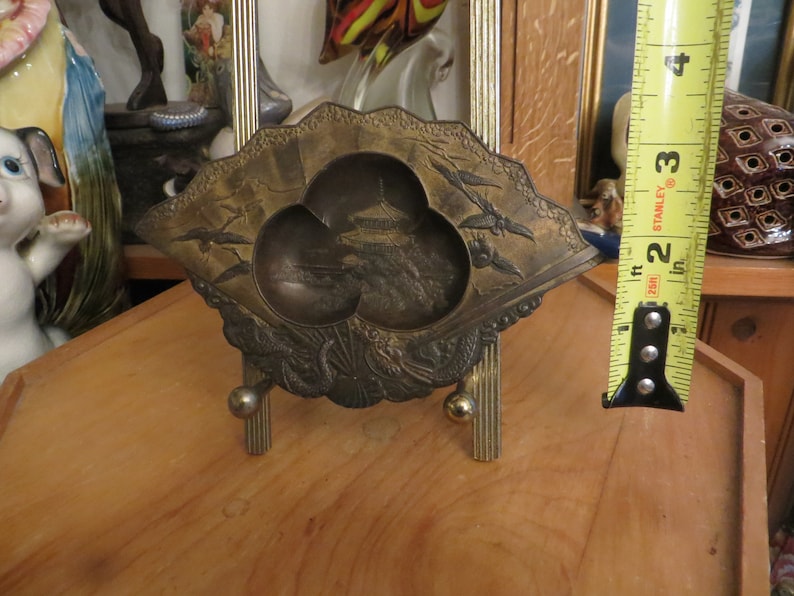 May include: A decorative brass tray with a fan-shaped design. The tray features a raised, three-dimensional image of a pagoda and a landscape. The tray is on a small, gold-coloured stand.
