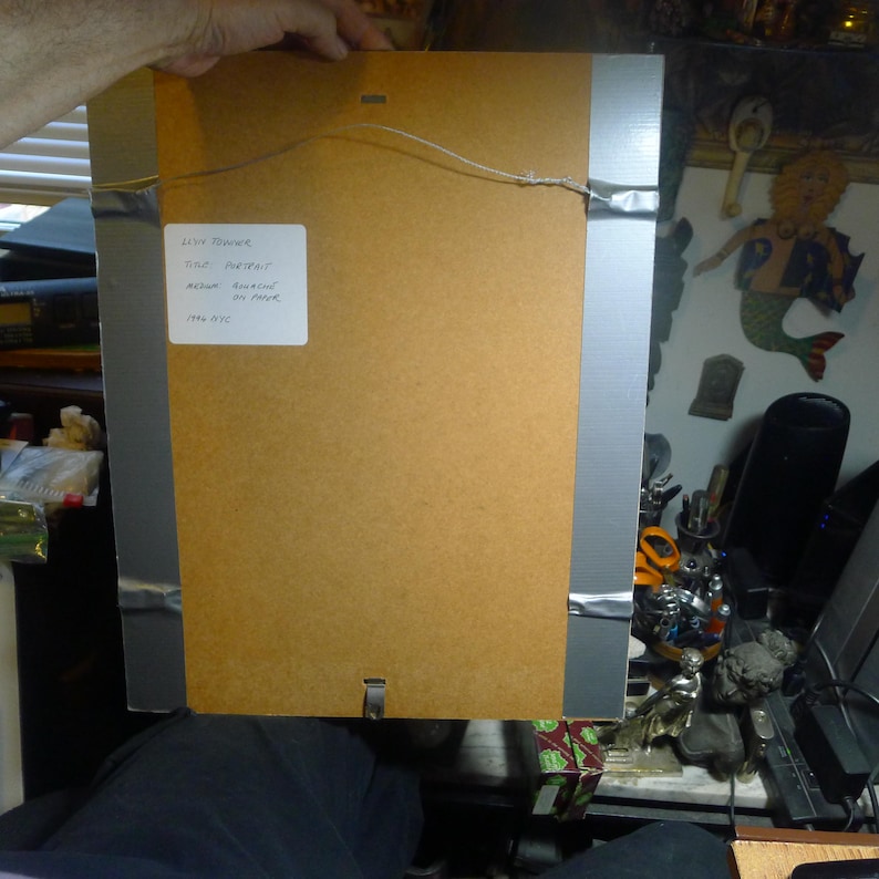 May include: Back of a framed artwork, with a brown cardboard backing and a silver metal frame. A white label with black text is affixed to the back. The artwork is held by a hand.