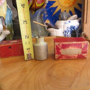 May include: A silver and white metal awl with a pointed tip, in a red cardboard box. The awl is about 2 inches long and is sitting on a wooden surface. A yellow tape measure is visible in the background.