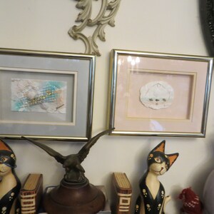 May include: Two framed pieces of art with gold frames, one with a light blue background and the other with a pink background.  The art pieces are displayed on a wall with a decorative mirror and a vase with flowers.
