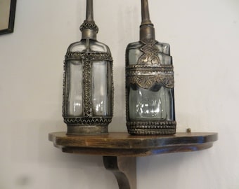 May include: Two antique glass bottles with ornate metal detailing, sitting on a wooden shelf. The bottles are clear glass with silver and gold accents.