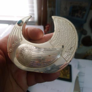 May include: A silver-toned bird-shaped brooch with intricate etched details. The brooch features a turquoise-colored stone and a detailed wing design. The bird's body has a floral pattern. The brooch is held in a hand.