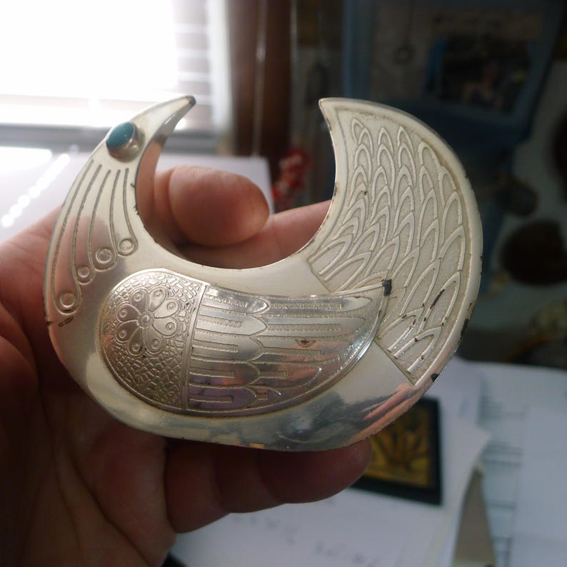 May include: A silver-toned bird-shaped brooch with intricate etched details. The brooch features a turquoise-colored stone and a detailed wing design. The bird's body has a floral pattern. The brooch is held in a hand.