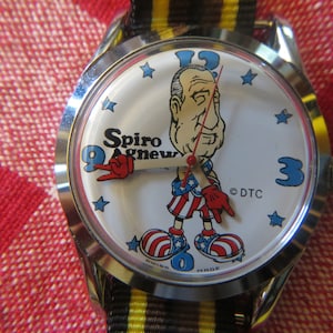 May include: A white wristwatch with a cartoon image of Spiro Agnew wearing a red, white, and blue outfit. The watch face has black Roman numeral hour markers and black hands. The watch band is black and yellow striped.
