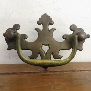 May include: Antique brass drawer pull with a decorative, ornate design. The pull has a curved handle and a center piece with a cutout.