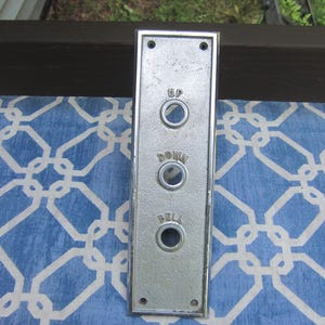 May include: A rectangular, silver-colored elevator button panel with three circular buttons. The panel has the words "UP", "DOWN", and "BELL" above each button. The panel is slightly weathered and has four small screw holes.