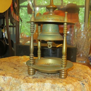 May include: An antique brass bell with a green patina, mounted on a decorative stand. The bell features intricate etched designs and is supported by four ornate legs. The stand includes a circular base and a top with a decorative finial.