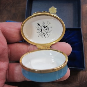 May include: A small, round, blue and white trinket box with a gold trim. The lid features a black and white illustration of musical instruments.
