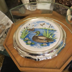 Puede incluir: Un soporte para tartas plateado con una parte superior de cer&aacute;mica blanca con un dise&ntilde;o de pato azul y verde y nen&uacute;fares.