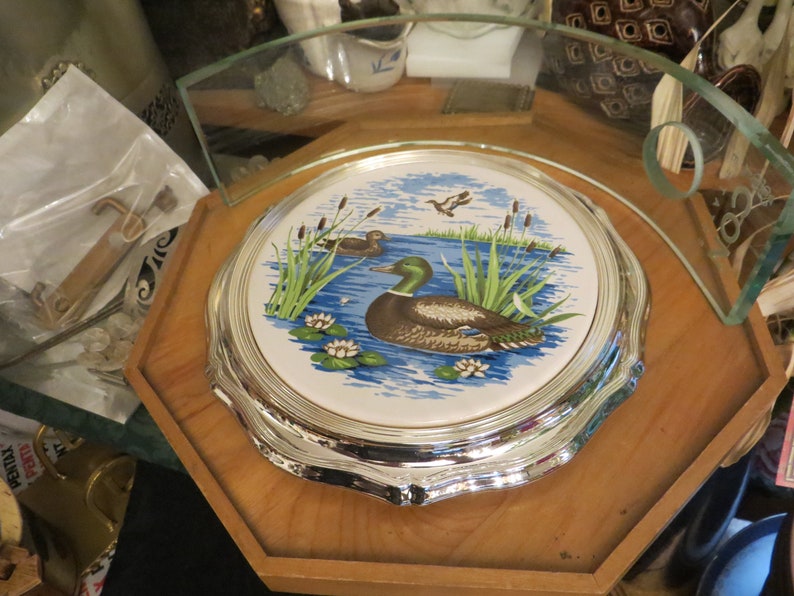 Puede incluir: Un soporte para tartas plateado con una parte superior de cer&aacute;mica blanca con un dise&ntilde;o de pato azul y verde y nen&uacute;fares.