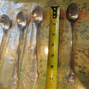 May include: Set of six silver teaspoons with ornate handles, packaged in plastic, next to a single silver teaspoon with an ornate handle. The spoons are 7 inches long.