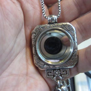 Pu&ograve; includere: Una collana con pendente argentato con cornice quadrata e volute decorative. Il pendente presenta un design circolare con un centro scuro e una superficie riflettente. La collana &egrave; tenuta in una mano.