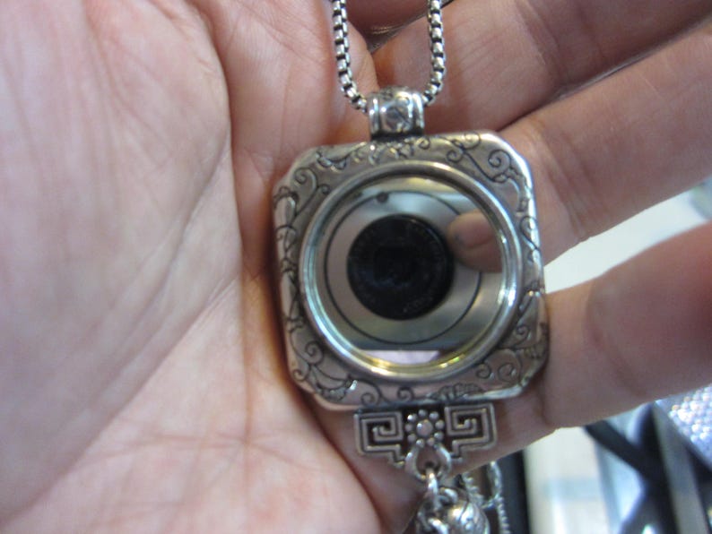 Pu&ograve; includere: Una collana con pendente argentato con cornice quadrata e volute decorative. Il pendente presenta un design circolare con un centro scuro e una superficie riflettente. La collana &egrave; tenuta in una mano.