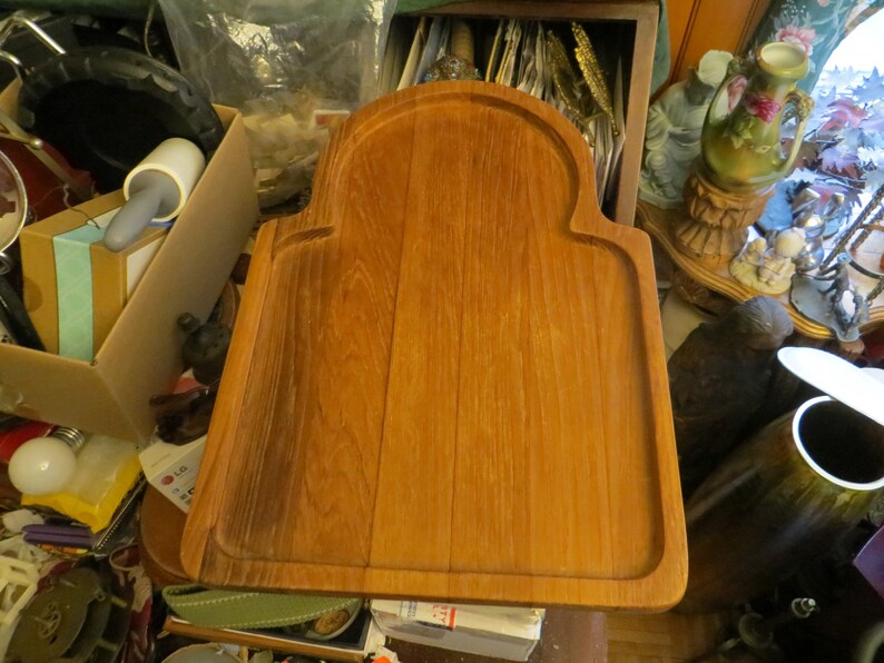 May include: A large, rectangular wooden serving board with a raised edge and a rounded arch at the top. The board is made of light brown wood with a natural grain pattern.