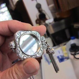 Pu&ograve; includere: Un specchietto compatto quadrato argentato con volute ornate e una piccola catena. Lo specchietto ha un nappa decorativa e un'apertura a cerniera. Il compatto &egrave; tenuto in una mano.