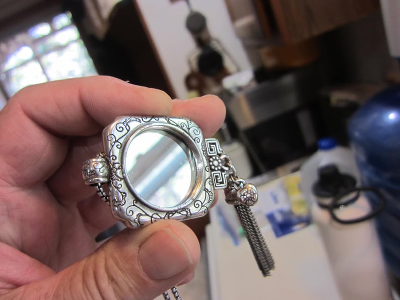 Pu&ograve; includere: Un specchietto compatto quadrato argentato con volute ornate e una piccola catena. Lo specchietto ha un nappa decorativa e un'apertura a cerniera. Il compatto &egrave; tenuto in una mano.