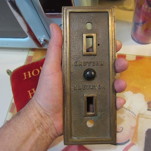 May include: A brass elevator button with the text "Eastern Elevator" and a black button in the center.