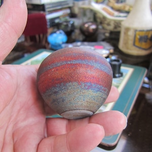 May include: A small, round, ceramic vase with a blue, gold, and red striped pattern. The vase is hand-painted and has a rustic, earthy look.