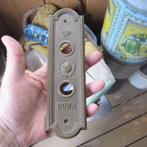 May include: A vintage brass elevator call button panel. The rectangular panel has three circular button openings and is engraved with the words "UP" and "DOWN". A decorative emblem is centered between the buttons.