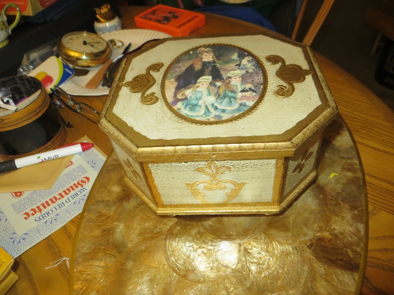 May include: Octagonal wooden box with a gold trim and a painted scene of three girls in a park. The box has a white background with gold accents.