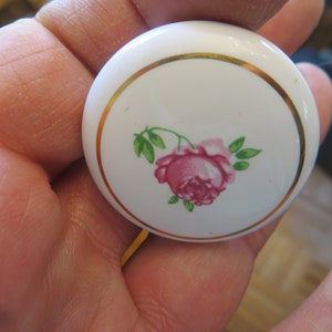 May include: A white ceramic cabinet knob with a pink rose design. The knob has a gold-colored trim around the edge of the floral design. The rose has green leaves and stems.