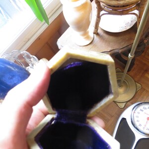 May include: A small, octagonal, beige ring box with a purple velvet interior. The box is open and the lid is resting on the side.