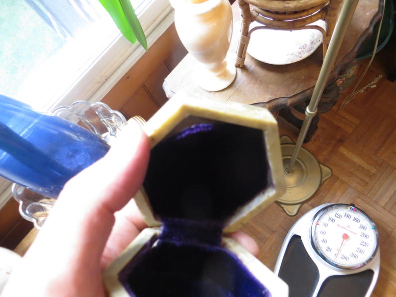 May include: A small, octagonal, beige ring box with a purple velvet interior. The box is open and the lid is resting on the side.