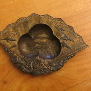 May include: A small, fan-shaped, brass trinket dish with three compartments. The dish is decorated with a pagoda, birds, and dragons.