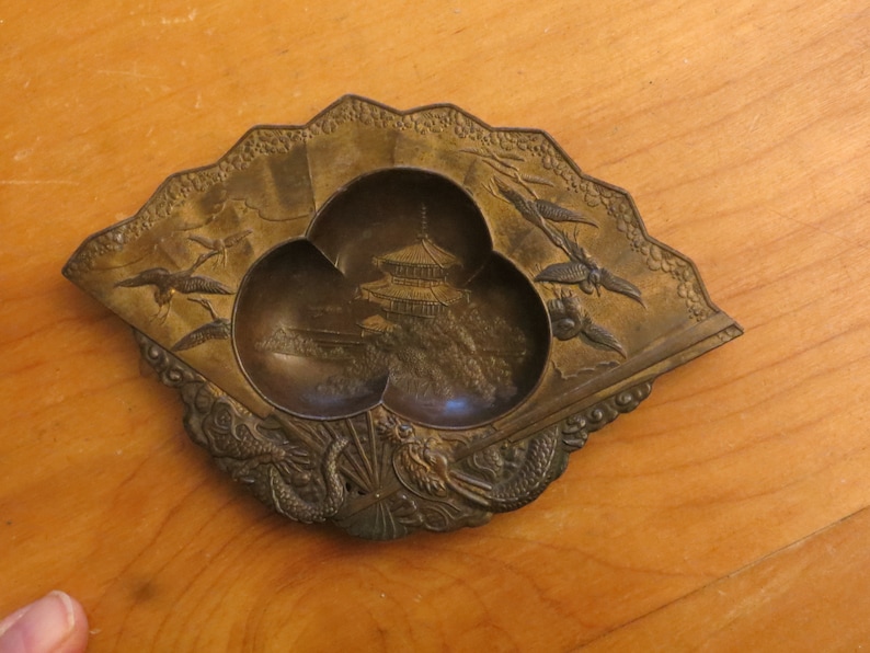 May include: A small, fan-shaped, brass trinket dish with three compartments. The dish is decorated with a pagoda, birds, and dragons.