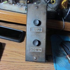 May include: A rectangular, metal elevator control panel with two round buttons and the words "UP" and "DOWN" engraved. The panel is a weathered, aged bronze color. The buttons are dark gray.